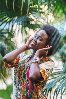 Fröhliche afrikanische Frau in farbenfroher traditioneller Kleidung lächelt und hört Musik über Kopfhörer, während sie inmitten von Palmenblättern steht - ADSF12264