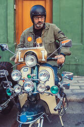 Bearded mature man in helmet and glasses sitting on custom motorcycle with many headlights on city street - ADSF12268