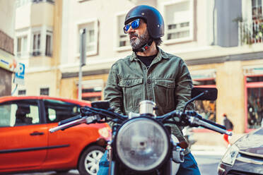 Serious bearded man in sunglasses and helmet sitting on motorbike during ride on city street looking away - ADSF12269