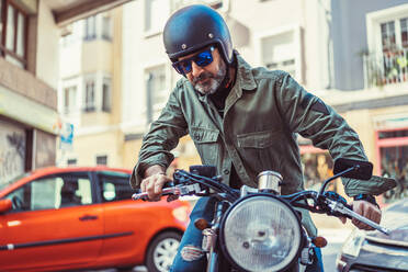 Ernster bärtiger Mann mit Sonnenbrille und Helm sitzt auf einem Motorrad während der Fahrt auf einer Stadtstraße und schaut weg - ADSF12270