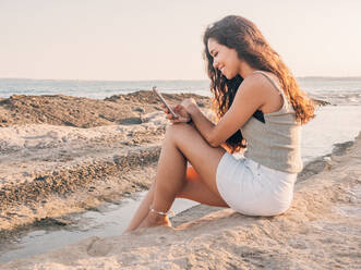 Verträumte trendige junge ethnische Frau, die am Strand SMS schreibt - ADSF12277