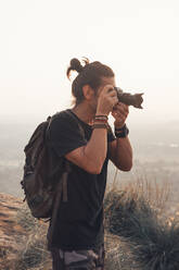 Side view of young male traveler in casual clothes with haversack standing on edge of cliff and taking picture of majestic landscape of green forest during sunset - ADSF12284