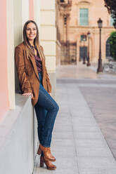 Satisfied charming lady in stylish casual clothing and heels leaning on wall of building and looking away at city street - ADSF12297