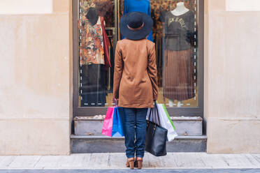 Rückenansicht einer nicht erkennbaren Dame mit modischem Hut und brauner Jacke, die durch ein Schaufenster auf ein modisches Kleid mit Rosen in einer Stadtstraße schaut - ADSF12304