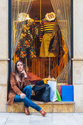 Stylish langhaarige weibliche Shopaholic in lässigen modische Kleidung sitzen mit Einkaufstaschen auf geschaukelt Zaun von Schaufenster, während lächelnd und Blick weg auf Stadt Straße - ADSF12305