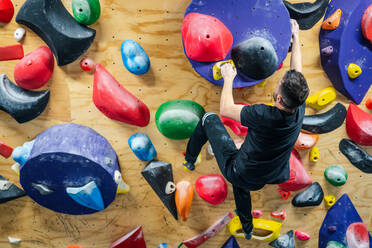 Rückenansicht eines nicht erkennbaren starken männlichen Sportlers in Sportkleidung, der während des Trainings in einem modernen Kletterzentrum an einer bunten Wand klettert - ADSF12323