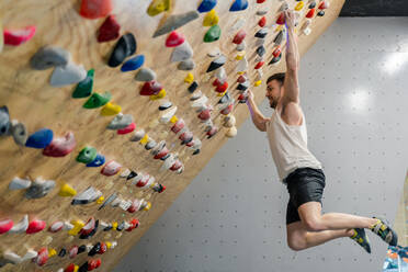 Side view from below of young man in active wear holding grips tightly while hanging in air during climbing training in gym - ADSF12332