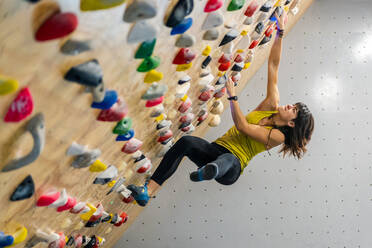 Seitenansicht von unten einer fokussierten Frau in Sportkleidung, die an einer steilen Wand über Matten in einem modernen Kletterzentrum hängt - ADSF12333