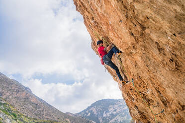 From below faceless sportive male alpinist climbing on sheer cliff in summer day - ADSF12340