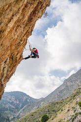 From bellow side view of sportive female alpinist ascending on cliff in mountain terrain in cloudy day - ADSF12341