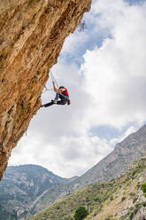 Von unten Seitenansicht einer sportlichen Bergsteigerin beim Aufstieg auf einen Felsen im Gebirge an einem bewölkten Tag - ADSF12341