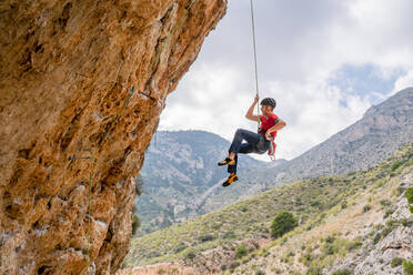 From bellow side view of sportive female alpinist ascending on cliff in mountain terrain in cloudy day - ADSF12342