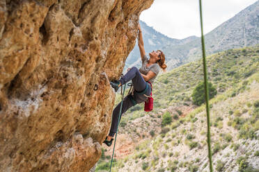 Fearless young female alpinist ascending on vertical cliff in mountain location in summertime - ADSF12345