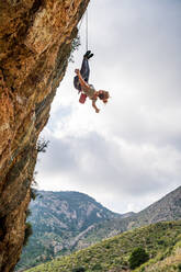 Unerschrockene junge Bergsteigerin genießt das Klettern und das Adrenalin, während sie kopfüber am Felsen hängt - ADSF12346