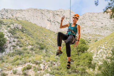 Fröhliche, sportliche Bergsteigerin, die in die Kamera schaut, während sie an einer Klippe unter bewölktem Himmel hängt - ADSF12349