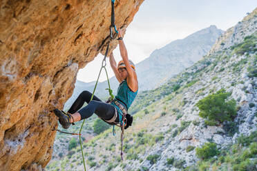 From below side view of active focused youthful female alpinist climbing on cliff in summer day - ADSF12352