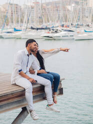 Young lovely couple sitting on dock with feet and embracing looking along - ADSF12371