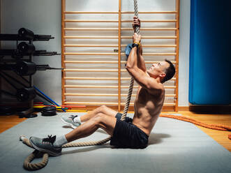 Seitenansicht des hemdlosen muskulösen Sportlers, der beim Training in einem modernen Fitnessstudio ein Seil hochklettert - ADSF12379