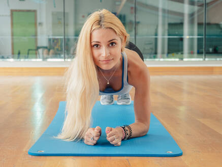 Schlanke Frau in Sportkleidung schaut in die Kamera, während sie auf einer Matte im Fitnessstudio eine Planke macht - ADSF12384