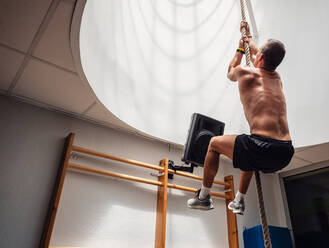 Rückenansicht eines unkenntlichen, hemdlosen, muskulösen Sportlers, der beim Training in einem modernen Fitnessstudio ein Seil hochklettert - ADSF12405