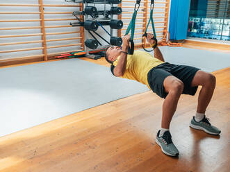 Erwachsener Mann in Sportkleidung hängt an TRX-Seilen und hält den Körper parallel zum Boden, während er in einem modernen Fitnessstudio trainiert - ADSF12408