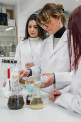 Mature woman in white coat showing chemical experiment with liquid to young female trainees while working in contemporary lab - ADSF12430