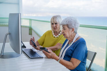 Stilvolle alte Freunde, die auf einem Tablet in den sozialen Medien surfen, während sie an einem Tisch mit Computer auf dem Balkon eines Resorts sitzen - ADSF12461