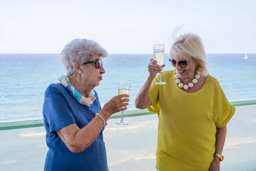 Glückliche reife Frauen in eleganten Outfits sagen einen Toast und genießen Wein, während sie auf dem Hotelbalkon in der Nähe des Meeres am Urlaubsort stehen - ADSF12465