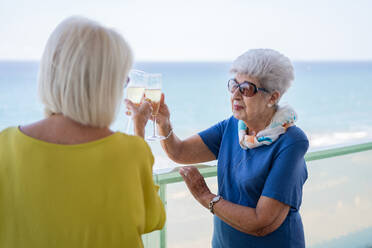 Glückliche reife Frauen in eleganten Outfits sagen einen Toast und genießen Wein, während sie auf dem Hotelbalkon in der Nähe des Meeres am Urlaubsort stehen - ADSF12466