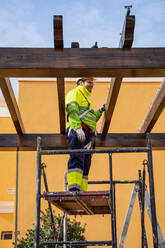 Von unten Seitenansicht eines männlichen Technikers in Arbeitskleidung, der auf einem Gerüst steht und sich auf die Installation eines Solarpanels auf einer Holzkonstruktion vorbereitet - ADSF12482