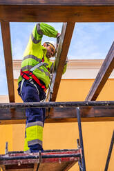 Von unten Seitenansicht eines männlichen Technikers in Arbeitskleidung, der auf einem Gerüst steht und sich auf die Installation eines Solarpanels auf einer Holzkonstruktion vorbereitet - ADSF12483