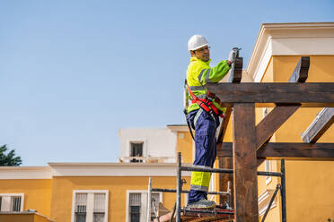 Von unten Seitenansicht eines männlichen Technikers in Arbeitskleidung, der auf einem Gerüst steht und sich auf die Installation eines Solarpanels auf einer Holzkonstruktion vorbereitet - ADSF12489