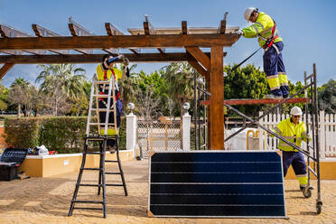 Gruppe von Arbeitern in Uniform und mit Helmen bei der Installation von Fotovoltaikanlagen auf dem Dach einer Holzkonstruktion in der Nähe eines Hauses - ADSF12492
