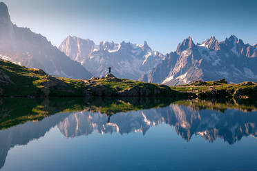 Lonely tourist on hilly shore reflecting in crystal lake in snowy mountains in sunlight - ADSF12511