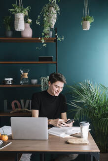 Lächelnder junger Mann, der sein Smartphone benutzt, während er mit Büchern und Laptop am Tisch sitzt - ALBF01379
