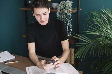 Gut aussehender junger Mann, der sein Smartphone benutzt, während er mit Büchern an einem Tisch vor einem Regal sitzt - ALBF01380