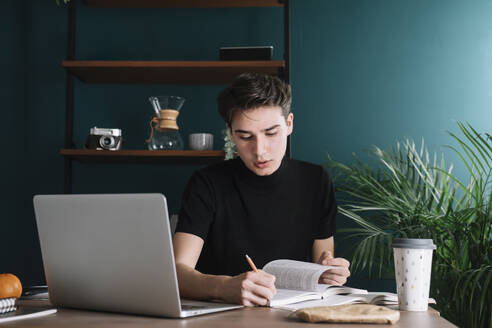 Männlicher Student, der in ein Buch schreibt, während er mit einem Laptop am Tisch sitzt und Hausaufgaben macht - ALBF01385