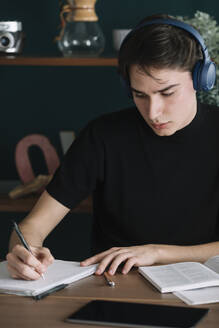 Hübscher junger Student, der in ein Buch schreibt, während er zu Hause über Kopfhörer Musik hört - ALBF01387
