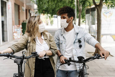 Couple wearing protective face masks while walking with electric bicycles on footpath - RDGF00130