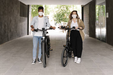 Couple in protective face masks standing with electric bicycles on footpath - RDGF00131