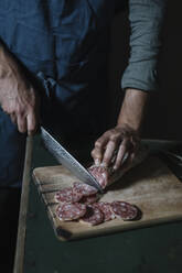 Midsection of man cutting salami slices on board at table - ALBF01410