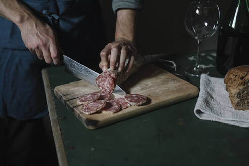 Hands of man cutting salami slices on board at table - ALBF01411
