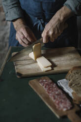 Mittelteil eines jungen Mannes, der handwerklich hergestellte Käsescheiben auf einem Brett am Tisch schneidet - ALBF01420