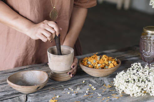 Midsection of woman crushing flowers and herbs in mortar with pestle on wooden table - ALBF01431