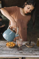 Schöne Frau gießt heißes Wasser in ein Glas und bereitet Kräutertee auf einem Holztisch zu - ALBF01440