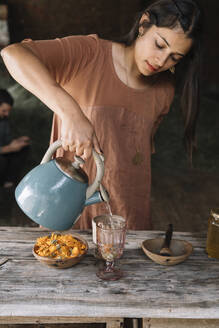 Beautiful woman pouring hot water in glass while preparing herbal tea on wooden table - ALBF01440