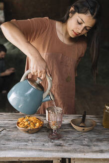 Schöne Frau gießt heißes Wasser in ein Glas und bereitet Kräutertee auf einem Holztisch zu - ALBF01440