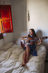 Junge Frau schaut weg, während sie zu Hause auf dem Bett Gitarre übt - LJF01758