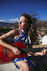 Schöne junge Frau mit geschlossenen Augen übt Gitarre auf dem Dach - LJF01764