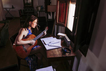 Young woman reading sheet music while practicing guitar at home - LJF01769